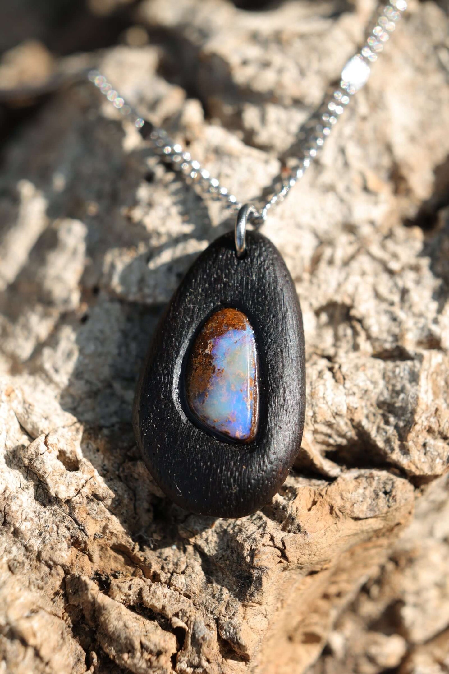 Holzkette - Boulder Opal in dunklem Edelholz mit elegantem Design aus Edelstahl auf natürlichem Untergrund.